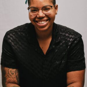 Smiling person with glasses, teal hair, tattoos